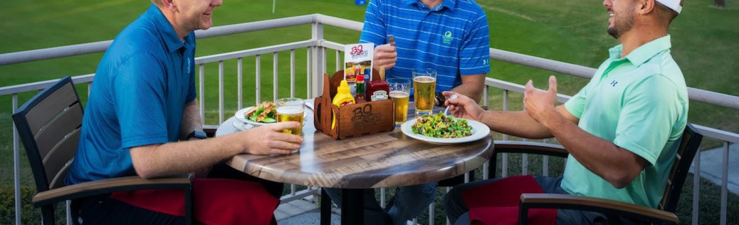 3 men in golf apparel laughing over drinks and food overlooking golf course