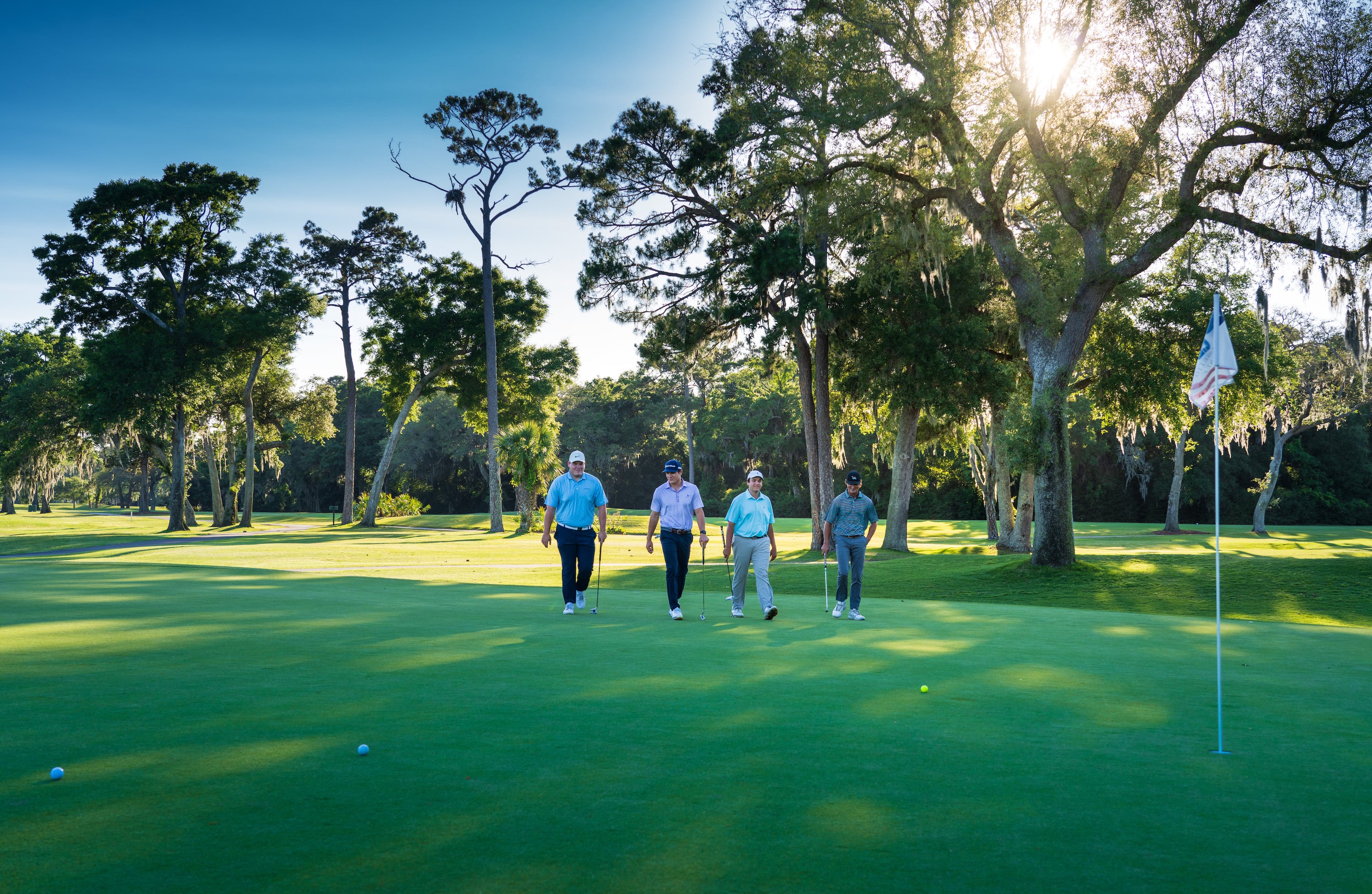 four male golfers walking towards golf balls and flag with sun and trees behind them